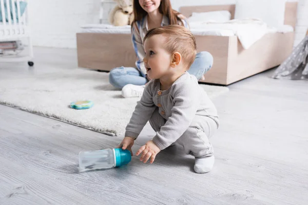 Niño feliz bebé sosteniendo biberón cerca de la madre sonriente sobre fondo borroso - foto de stock