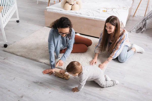 Vista ad alto angolo del bambino che gioca con auto giocattolo in legno vicino a donne multietniche felici — Foto stock