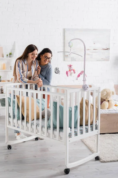 Heureux interracial femmes regarder bébé garçon dans bébé lit dans chambre — Photo de stock