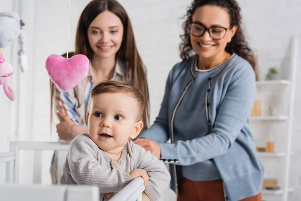 Femmes interraciales floues et heureuses regardant bébé garçon dans la crèche de bébé dans la chambre — Photo de stock