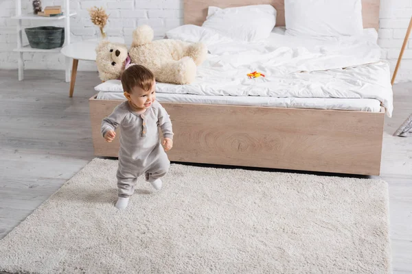 Happy baby boy in grey romper walking on carpet near soft toy on bed — Stock Photo