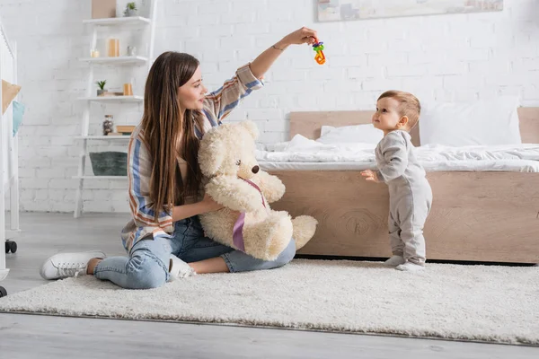 Erstaunte junge Mutter hält Teddybär und rasselt in die Nähe ihres kleinen Sohnes — Stockfoto