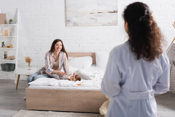 Back view of blurred african american pediatrician in white coat standing near happy mother with baby son on bed — Stock Photo