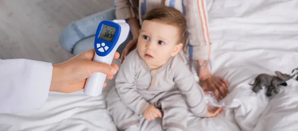 Arzt untersucht Säugling mit berührungslosem Thermometer in der Nähe der Mutter, Banner — Stockfoto