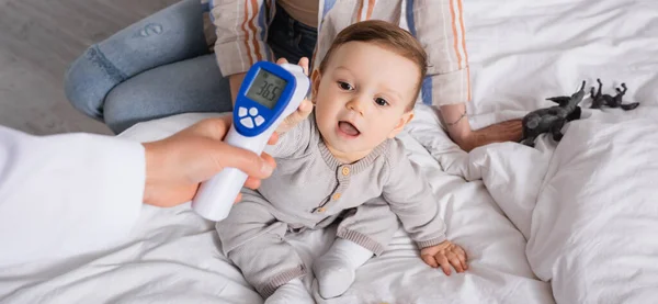 Pediatra examinando niño con termómetro sin contacto cerca de la madre, pancarta - foto de stock