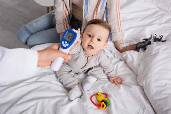Kinderarzt untersucht Jungen mit berührungslosem Thermometer in der Nähe der Mutter — Stockfoto