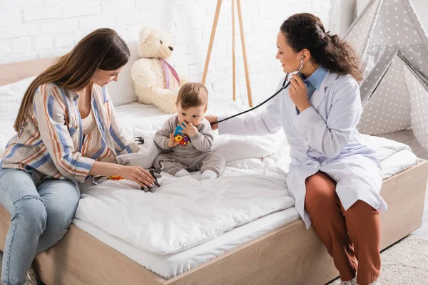 Smiling african american pediatrician in white coat sitting on bed and examining baby boy with stethoscope — Stock Photo