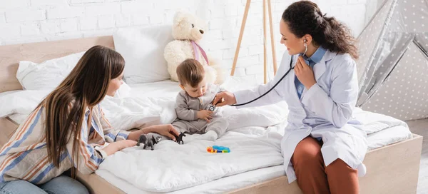 Happy african american pediatrician in white coat sitting on bed and examining baby boy with stethoscope, banner — Stock Photo