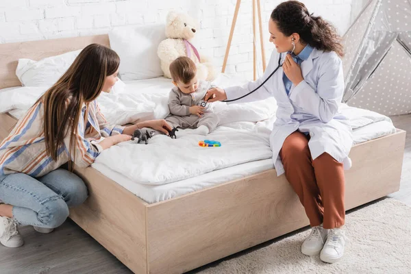 Happy african american pediatrician in white coat sitting on bed and examining baby boy with stethoscope — Stock Photo