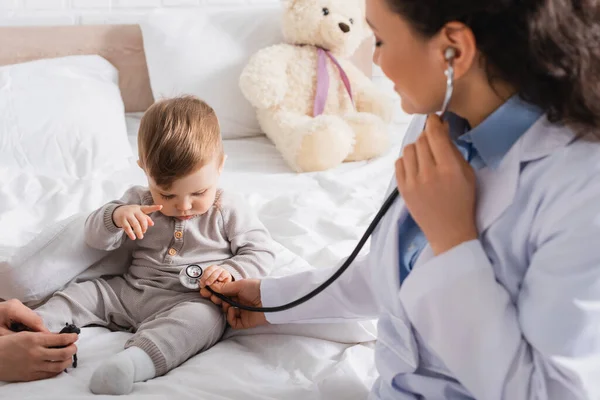 Pédiatre afro-américain flou en manteau blanc examinant bébé garçon avec stéthoscope près de la mère — Photo de stock