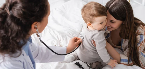 Pediatra afro-americano de casaco branco examinando menino com estetoscópio, bandeira — Fotografia de Stock