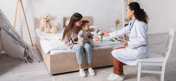Afrikanisch-amerikanischer Kinderarzt hält digitales Tablet in der Hand und gibt Spielzeug an Säugling in der Nähe der Mutter, Banner — Stockfoto
