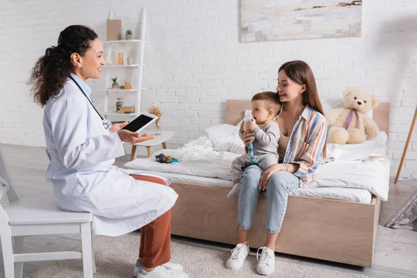 Glücklicher afrikanisch-amerikanischer Kinderarzt in weißem Mantel mit digitalem Tablet mit leerem Bildschirm in der Nähe von Mutter und Sohn im Schlafzimmer — Stockfoto