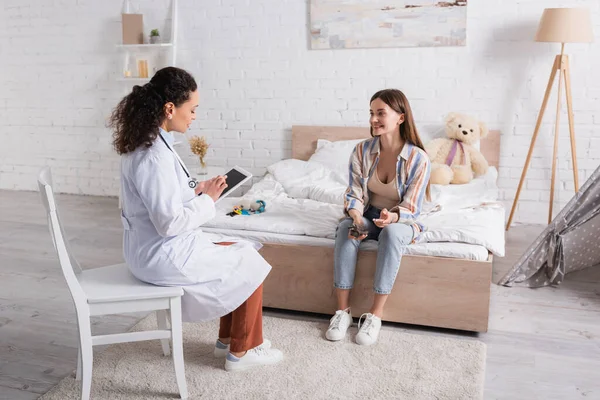 Médico afroamericano de capa blanca usando tableta digital cerca de mujer feliz sentada en el dormitorio - foto de stock