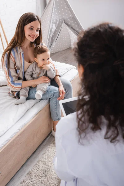 Glückliche Mutter, die mit ihrem Sohn sitzt und den afrikanisch-amerikanischen Arzt im verschwommenen Vordergrund betrachtet — Stockfoto