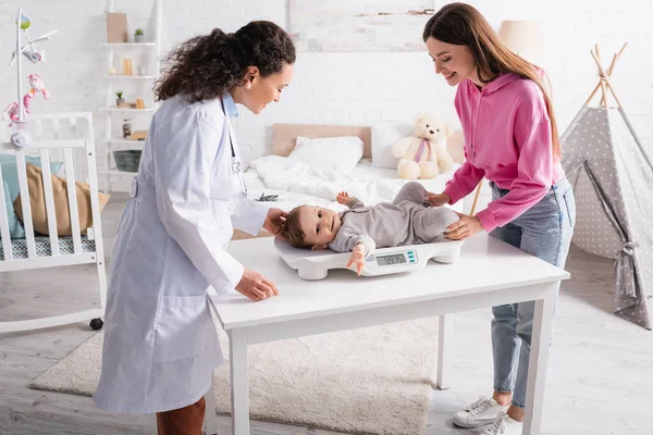 Happy african american pediatrician and mother looking at infant boy on baby scale — Stock Photo