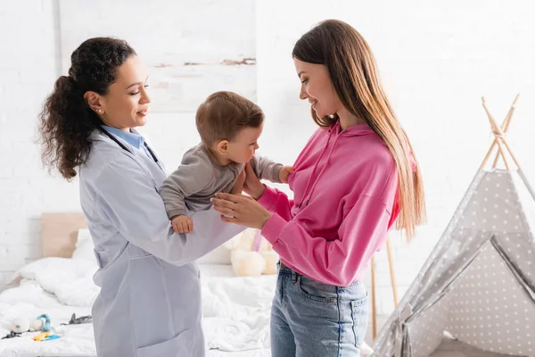 Lächelnder afrikanisch-amerikanischer Kinderarzt im weißen Mantel, der einen kleinen Jungen in der Nähe einer glücklichen Mutter hält — Stockfoto