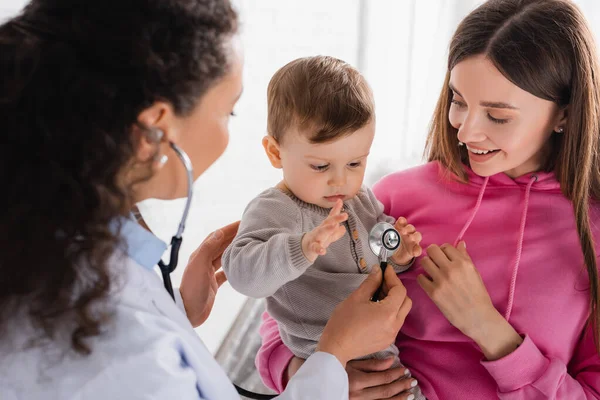 Offuscata africano americano pediatra esaminando bambino ragazzo in mani di madre sorridente — Foto stock