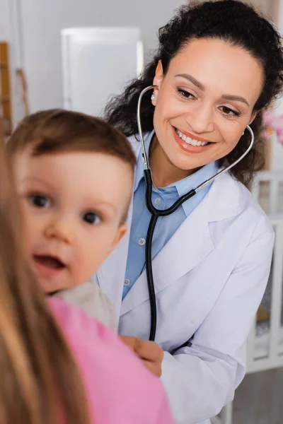 Happy african american pediatrician examining infant boy in hands of mother on blurred foreground — Stock Photo