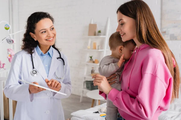 Glückliche Mutter hält kleinen Jungen in der Nähe von afrikanischem amerikanischen Kinderarzt in den Armen und zeigt auf digitales Tablet — Stockfoto