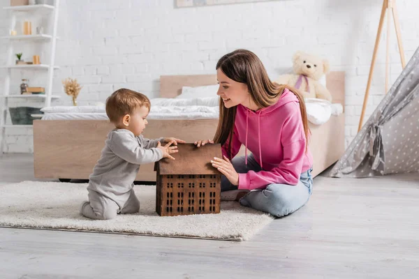 Happy mother looking at infant son sitting on carpet near toy house — Stock Photo