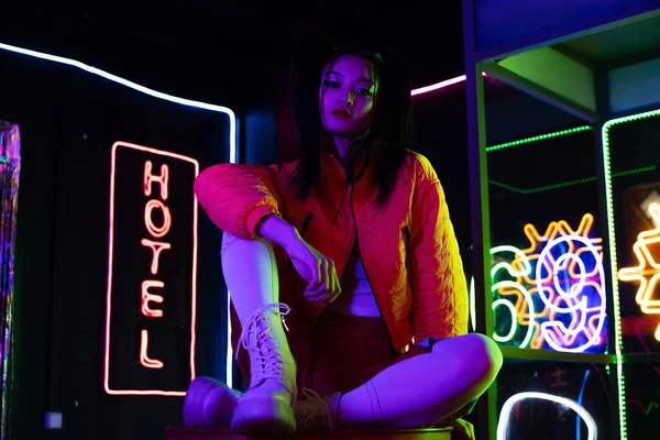 Brunette young asian woman sitting near neon sign with hotel lettering outside — Photo de stock