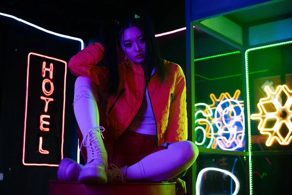 Trendy young asian woman sitting near neon sign with hotel lettering outside - foto de stock