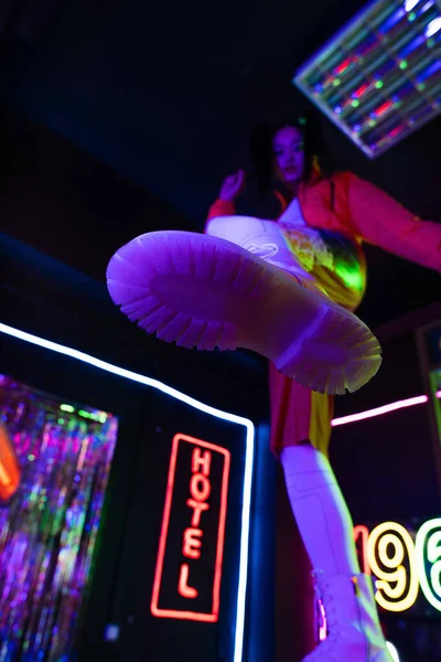 Low angle view of blurred young asian woman in white shoes near neon sign with hotel lettering outside — Stock Photo