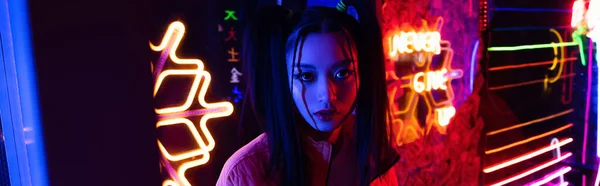 Stylish young asian woman looking at camera near neon lighting outside, banner — Foto stock