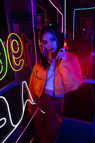 Neon lighting on stylish young asian woman talking on retro payphone outside — Stock Photo