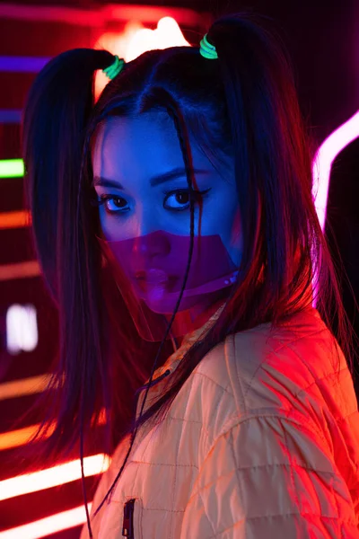 Trendige junge Asiatin mit Gesichtsschutz in der Nähe von Neonbeleuchtung — Stockfoto