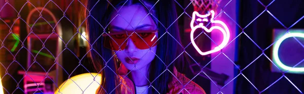 Trendy young asian woman in red sunglasses near neon sign and metallic fence, banner — Foto stock
