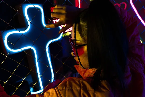 Stylish young asian woman in sunglasses posing near neon sign and metallic fence - foto de stock