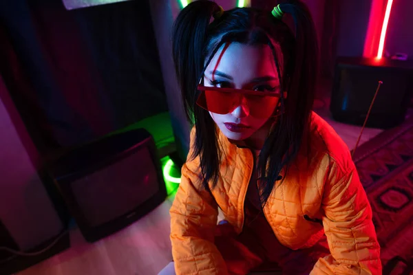 High angle view of stylish young asian woman in sunglasses and orange jacket posing near neon lighting — Foto stock