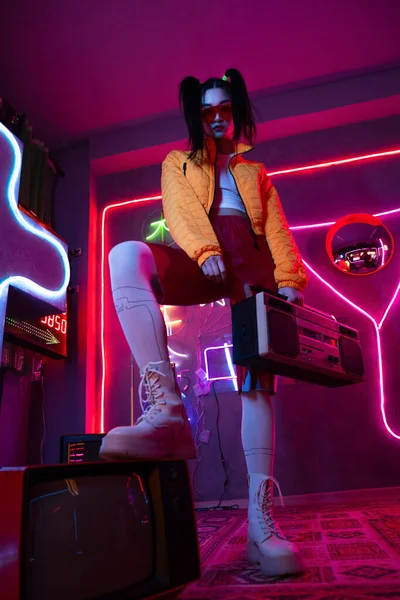 Low angle view of young asian woman in sunglasses and orange jacket posing with retro boombox near tv - foto de stock