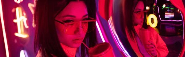 Stylish young asian woman in sunglasses posing with paper cup near mirror and neon lighting, banner — Stock Photo