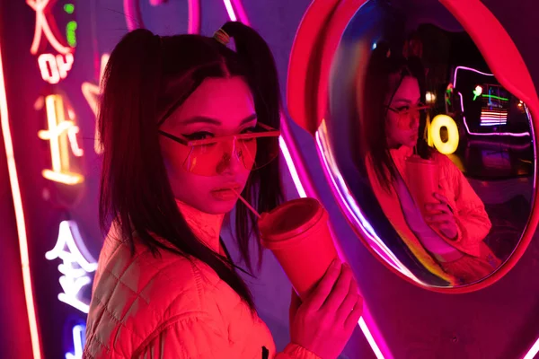 Stylish asian woman in sunglasses posing with paper cup near mirror and neon lighting — Fotografia de Stock