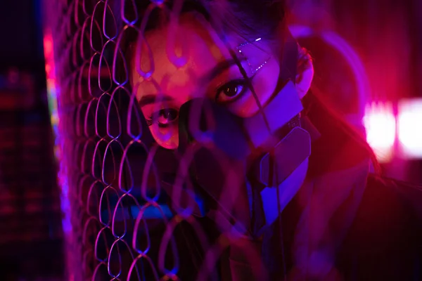 Neon lighting on young asian woman in gas mask looking through metallic fence — Photo de stock