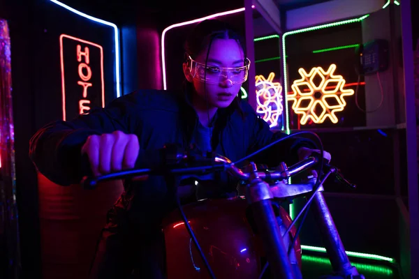 Young asian woman in sunglasses riding motorcycle near neon sign outside — Stock Photo