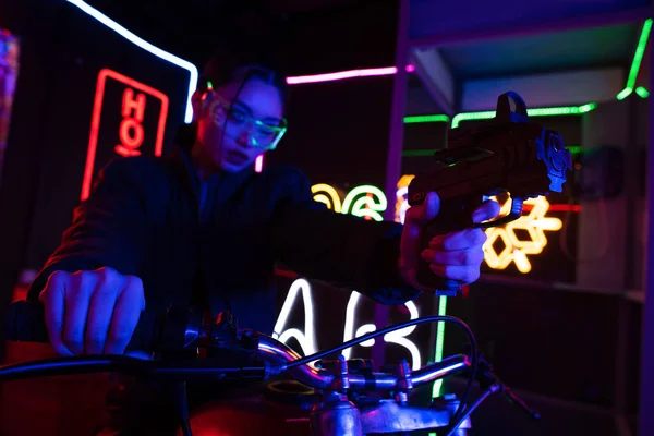 Blurred asian woman in sunglasses aiming with gun while riding motorcycle near neon sign — Fotografia de Stock