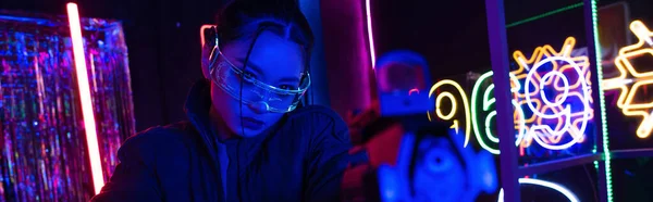 Armed asian woman in sunglasses aiming with gun near neon sign, banner — Stock Photo