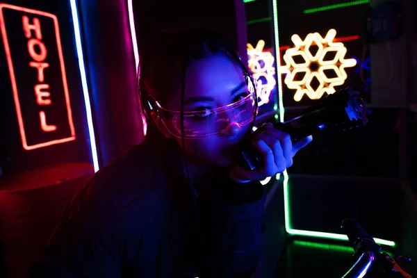 Bewaffnete Asiatin mit Sonnenbrille hält Waffe in der Nähe von neonfarbenem Hotelschild — Stockfoto