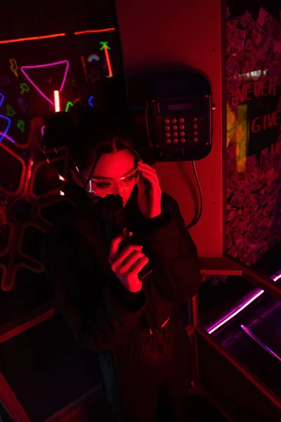 Armed asian woman in sunglasses holding gun while talking on retro payphone — Foto stock