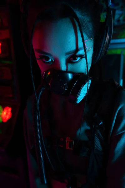Young asian woman with scar in gas mask and wireless headphones looking at camera — Fotografia de Stock