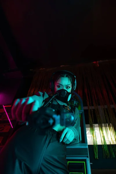 Low angle view of armed asian woman in gas mask and wireless headphones looking at camera while aiming with gun — Stock Photo