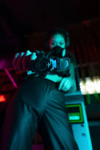 Low angle view of blurred asian woman in gas mask and wireless headphones looking at camera while aiming with gun - foto de stock