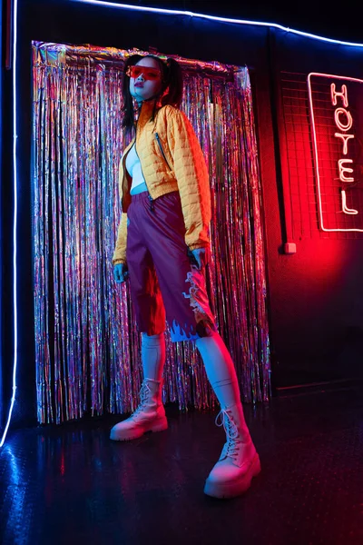 Full length of young asian woman in sunglasses looking at camera near red neon hotel sign — Stock Photo