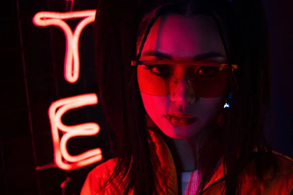 Освещение молодой азиатской женщины в солнечных очках, смотрящей на камеру рядом с красным неоновым знаком — стоковое фото