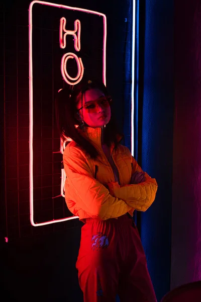 Young asian woman in sunglasses standing with crossed arms near red neon hotel sign — Stock Photo