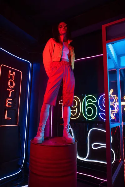 Full length of asian woman in sunglasses looking at camera while posing near red neon hotel sign - foto de stock